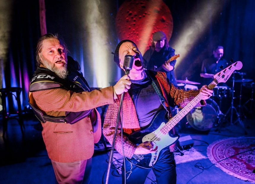 Olli-Kalle Heimo ojentaa mikkiä kitaraa soittavalle Jaakko Tohkaselle. Bändi soittaa takana.