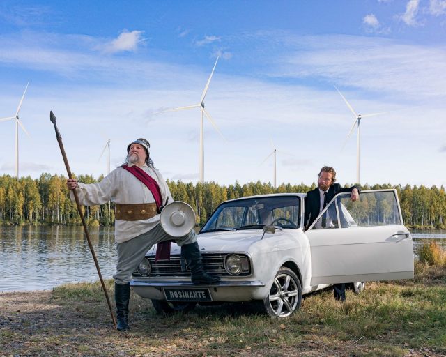 Olli-Kalle Heimo on pukeutunut ritarilliseen asuun ja poseeraa keihäs kädessään vanhan auton vieressä. Jaakko Tohkanen katsoo ihmeissään puku päällä. Jokimaisemassa tuulivoimaloita.