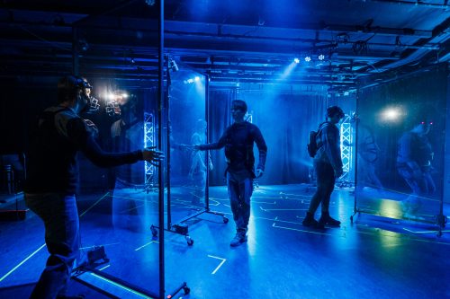 Ihmishahmoja liikuskelee sinisellä valolla valaistuneessa tilassa, jossa on heijastavia ja läpi näkyviä lasiseiniä.
