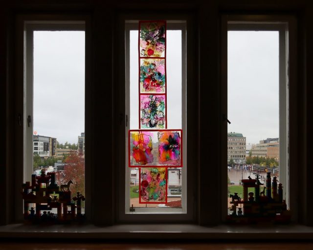 Kapeiden korkeiden ikkunoiden eteen on aseteltu kaksi pienempää rakennelmaa ja keskimmäiseen ikkunaan läpikuultavia, värikkäitä teoksia. Taustalla näkyy Joensuun keskustaa.