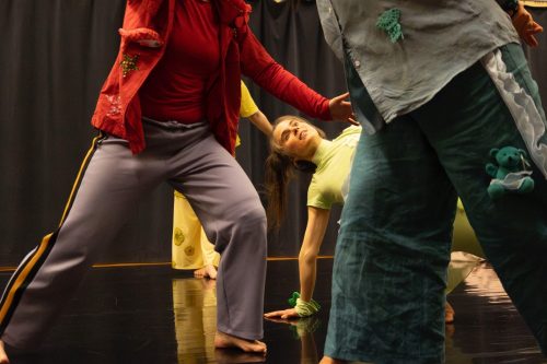 Lähikuva tanssijasta, joka nojaa käsillään lattiaan. Etualalla liikeepätarkkoja tanssijoita.