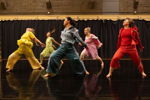 Viisi värikkäästi pukeutunutta tanssijaa ottaa jättiaskeleen vasemmalle suunnaten katseensa menosuuntaan.