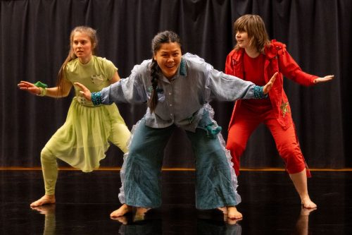 Tanssija edessä on lkyyryssä ja eveässä haara-asennossa kädet levitettyinä- Hän katsoo kameraan hymyillen.