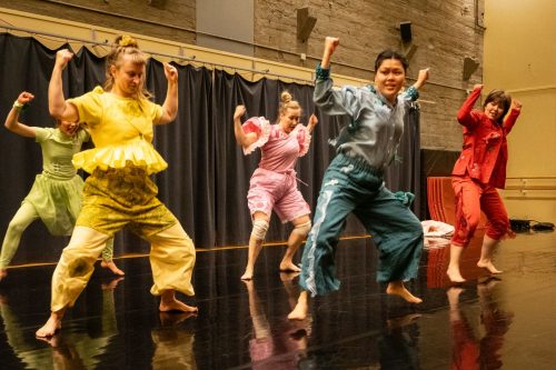 Joukko värikkäisiin pukeutuneita tanssijoita ovat asennossa, jossa jalat ovat harallaan ja kädet ylösnostettuina.