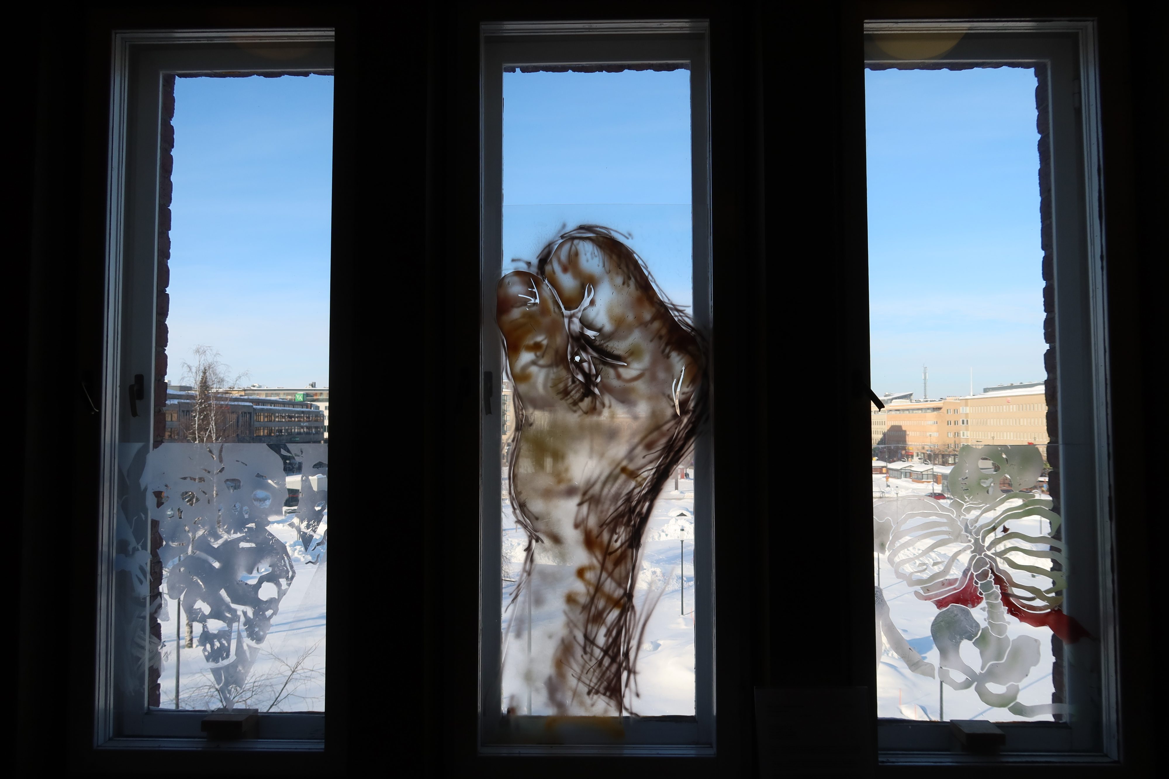 Kolme läpinäkyvää taideteosta ikkunoissa. Taustalla näkyy aurinkoinen talvimaisema.