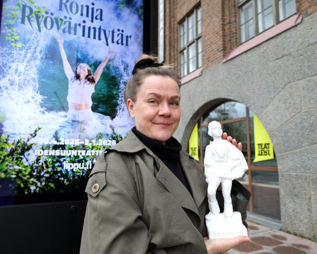 Nainen esittelee kipsipatsasta hymyillen Ronja Ryövärintyttären julisteen edessä.