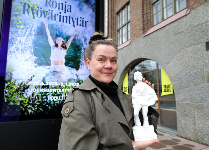 Nainen esittelee kipsipatsasta hymyillen Ronja Ryövärintyttären julisteen edessä.