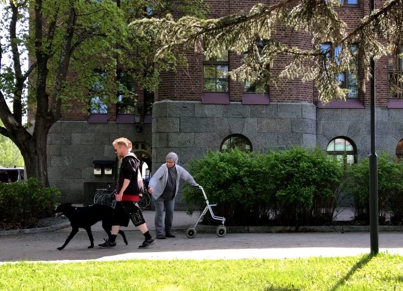 Aimo Kätkäläinen (Regina Launivuo) seisoo kadulla rollaattoriin nojaten ja ojentaa kättään kohti vastaan tulevaa nuoren miehen taluttamaa mustaa koiraa.