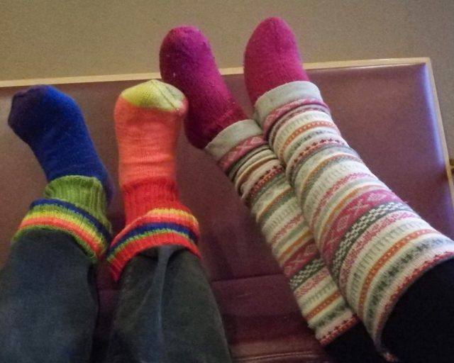 Kahdet jalat on oikaistu penkille. Molemmilla henkilöillä on jalassaan värikkäät, raidalliset villasukat.